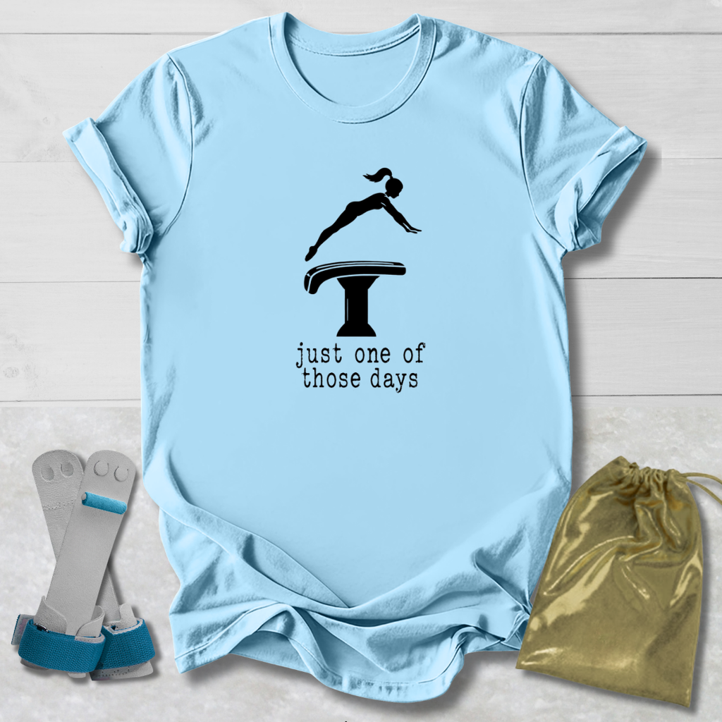 A light blue graphic T-shirt laid flat on a white wood background, featuring a black gymnast silhouette on a vaulting table with the text 'just one of those days.' A pair of gymnastics straps and a gold drawstring bag are placed beside the shirt as styling props.