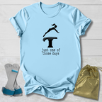 A light blue graphic T-shirt laid flat on a white wood background, featuring a black gymnast silhouette on a vaulting table with the text 'just one of those days.' A pair of gymnastics straps and a gold drawstring bag are placed beside the shirt as styling props.