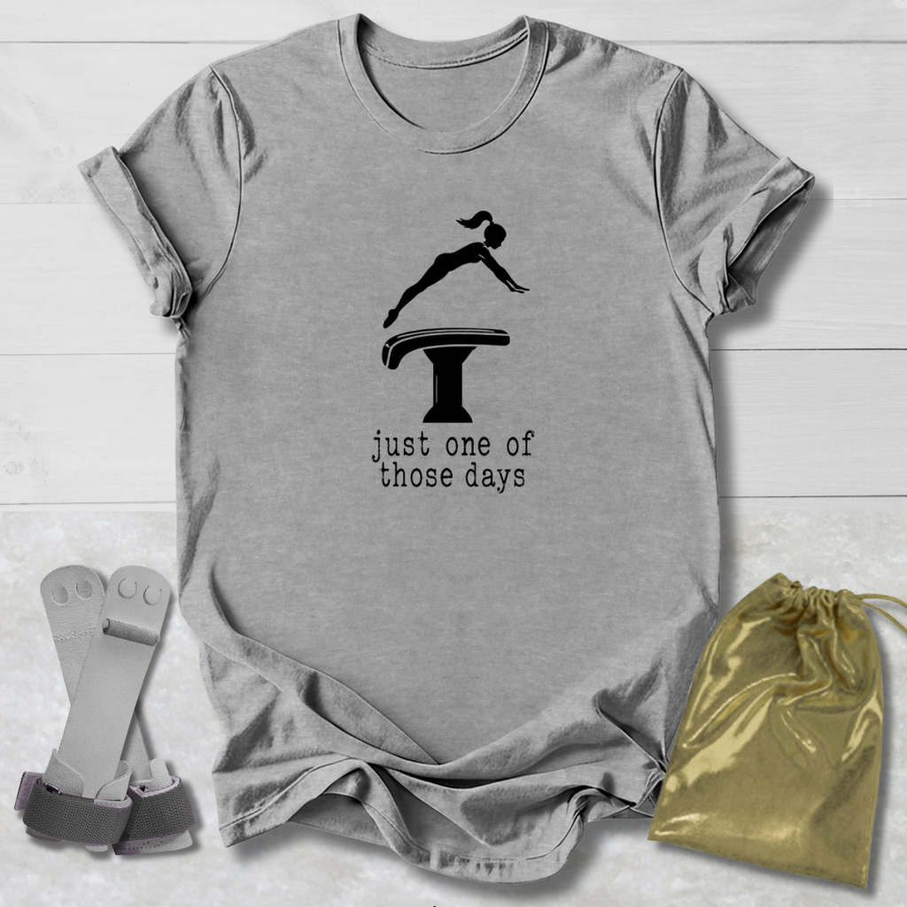 A heather gray short-sleeve t-shirt laid flat on a white wood background, featuring a black graphic of a gymnast on a high jump table with the text 'just one of those days'. A gold drawstring pouch and a pair of gray athletic straps are placed beside the shirt.