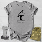 A heather gray short-sleeve t-shirt laid flat on a white wood background, featuring a black graphic of a gymnast on a high jump table with the text 'just one of those days'. A gold drawstring pouch and a pair of gray athletic straps are placed beside the shirt.