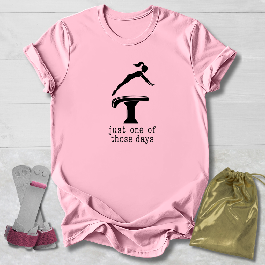 A light pink short-sleeve T-shirt laid flat on a white wood background, featuring a black gymnast silhouette above a balance beam graphic with the text 'just one of those days.' Two gymnastics shoes are placed to the left, and a gold drawstring bag is to the right.