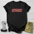 A black short-sleeve t-shirt laid flat on a light wood background, featuring the word 'GYMNAST' across the chest in leopard-print letters outlined in pink. A gold drawstring pouch and a pair of gray and black athletic straps are placed beside the shirt.