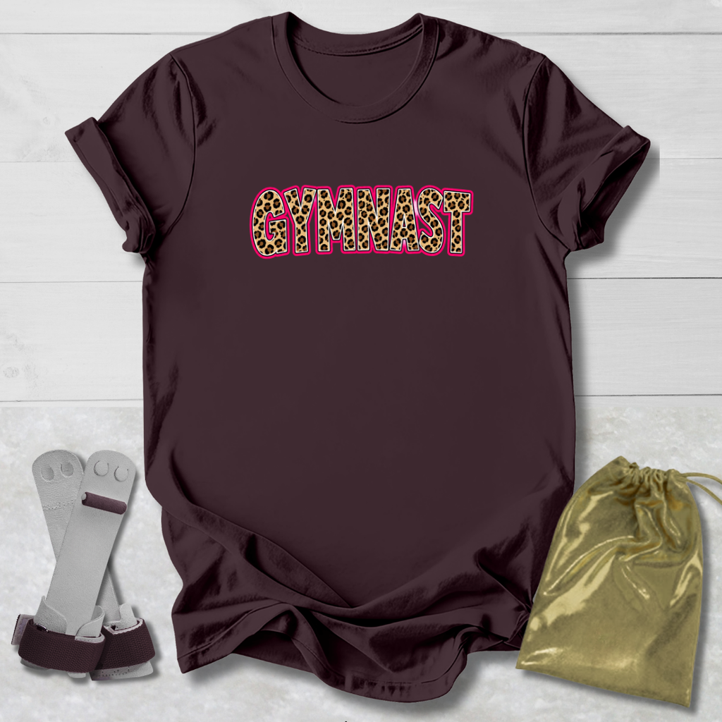 A dark brown short-sleeve t-shirt with the word 'GYMNAST' printed across the chest in a leopard-print pattern with a pink outline. The shirt is displayed on a light gray wood background alongside a pair of gymnastics bars and a gold drawstring bag.
