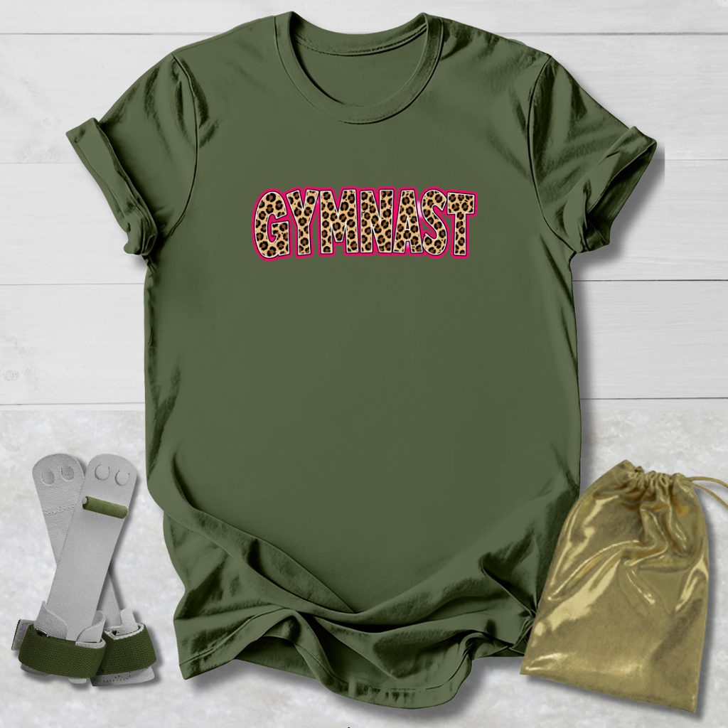 An olive green short-sleeve t-shirt with the word 'GYMNIST' printed across the chest in a pink-outlined leopard print. The shirt is displayed in a flat-lay arrangement on a white wood background, accompanied by a pair of white and green athletic straps and a gold drawstring bag.