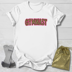 A white short-sleeve T-shirt laid flat on a light wood background, featuring the word 'GYMNAST' across the chest in a pink-outlined leopard-print pattern. A small gold drawstring pouch and gray athletic straps are placed beside the shirt as styling props.