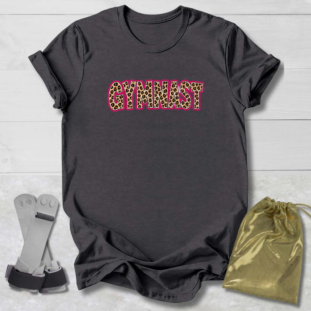A dark charcoal gray t-shirt with the word 'GYMNAST' printed across the chest in a leopard print pattern with a pink outline. The shirt is displayed flat on a light gray wood background, accompanied by a pair of gymnastics straps on the left and a gold drawstring bag on the right.
