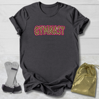 A dark charcoal gray t-shirt with the word 'GYMNAST' printed across the chest in a leopard print pattern with a pink outline. The shirt is displayed flat on a light gray wood background, accompanied by a pair of gymnastics straps on the left and a gold drawstring bag on the right.
