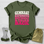 An olive green graphic t-shirt laid flat on a white wood background, featuring the text 'GYMNAST EXPENSIVE DIFFICULT & TALKS BACK' in white and pink bubble letters. A pair of white and green gymnastics straps and a gold drawstring pouch are placed beside the shirt as props.