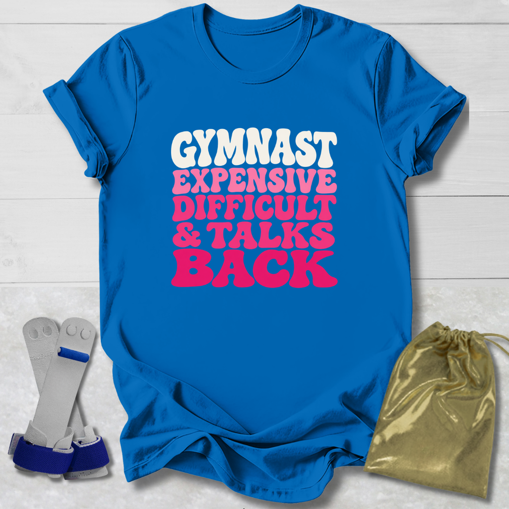A bright blue short-sleeve T-shirt laid flat on a white wood background, printed with the phrase 'GYMNAST EXPENSIVE DIFFICULT & TALKS BACK' in white and pink lettering. A gold drawstring bag and a pair of gray-and-blue gymnast wrist wraps are placed beside the shirt as styling props.