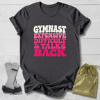 A dark charcoal gray graphic t-shirt laid flat on a white wood background, featuring the text 'GYMNAST EXPENSIVE DIFFICULT & TALKS BACK' in white and pink bubble lettering. A pair of gymnast straps and a shiny gold drawstring bag are styled as props next to the shirt.
