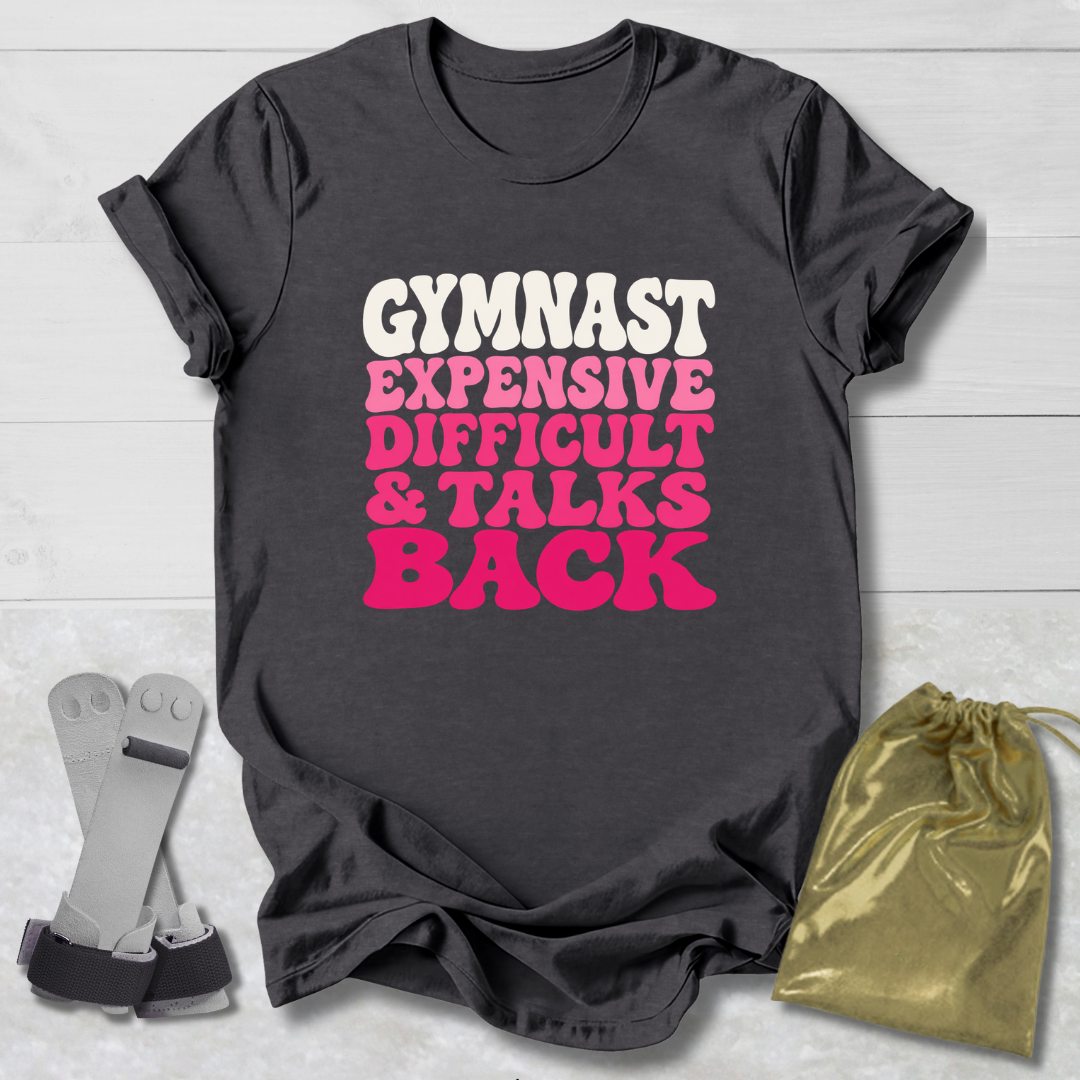 A dark charcoal gray graphic t-shirt laid flat on a white wood background, featuring the text 'GYMNAST EXPENSIVE DIFFICULT & TALKS BACK' in white and pink bubble lettering. A pair of gymnast straps and a shiny gold drawstring bag are styled as props next to the shirt.