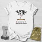 A white graphic t-shirt laid flat on a light wood background, featuring the text 'PRACTICE AT 8 A.M.