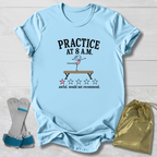 A light blue graphic t-shirt laid flat on a white wood background, featuring a gymnastics-themed print that reads 'PRACTICE AT 8 A.M.