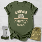 An olive green graphic t-shirt laid flat on a white wood background, featuring a cream-colored print that reads 'HOMEWORK PRACTICE REPEAT' with a stack of books and bench illustration. A pair of gray and green athletic wristbands and a gold drawstring pouch are placed beside the shirt.