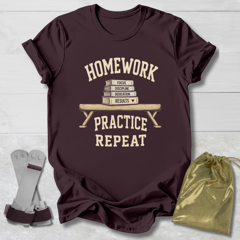 A dark maroon short-sleeve graphic T-shirt laid flat on a light wood background, with a cream print reading “HOMEWORK PRACTICE REPEAT” around an illustration of stacked books and a bench. A gray pair of dumbbells and a gold drawstring bag are placed beside the shirt as styling props.
