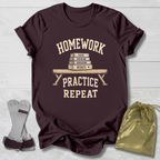 A dark maroon short-sleeve graphic T-shirt laid flat on a light wood background, with a cream print reading “HOMEWORK PRACTICE REPEAT” around an illustration of stacked books and a bench. A gray pair of dumbbells and a gold drawstring bag are placed beside the shirt as styling props.