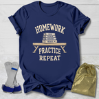 A navy blue graphic t-shirt laid flat on a white wood background, featuring the cream-colored text 'HOMEWORK PRACTICE REPEAT' with a stack of books and ruler illustration. A gold drawstring bag and gray/blue athletic straps are placed beside the shirt as styling props.