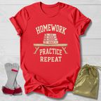A red short-sleeve graphic T-shirt laid flat on a light wood background, with a cream-colored school-themed print reading 'HOMEWORK PRACTICE REPEAT' around an illustration of stacked books on a bench. A gray and pink shoe accessory and a gold drawstring pouch are placed beside the shirt as styling props.