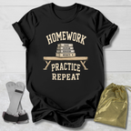 A black graphic t-shirt laid flat on a light wood background, featuring a cream-colored print that reads 'HOMEWORK PRACTICE REPEAT' with a stack of books and a pencil. A gray resistance band is placed to the left, and a shiny gold drawstring pouch is on the right.
