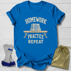 A bright blue graphic T-shirt laid flat on a white wood background, with cream text reading 'HOMEWORK PRACTICE REPEAT' and a stack of books on the front. Two small accessories are placed beside it: a pair of gray and blue wrist/ankle wraps on the left and a shiny gold drawstring pouch on the right.