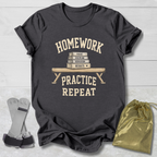 A dark charcoal gray graphic t-shirt laid flat on a white wood background, featuring a cream-colored print that reads 'HOMEWORK PRACTICE REPEAT' around an illustration of stacked books on a bench. A gold drawstring pouch and a pair of gray and black athletic shoes are placed beside the shirt as props.