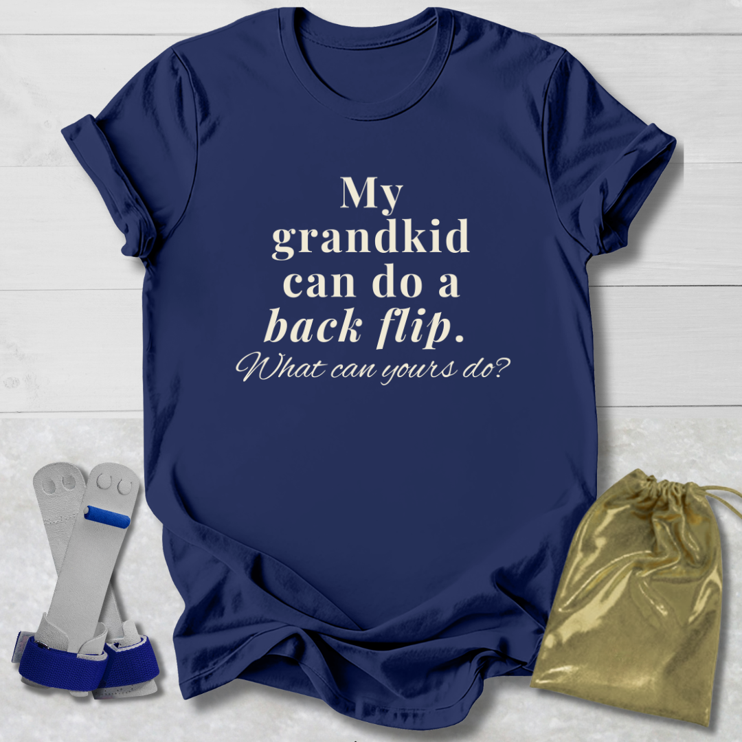A navy blue graphic t-shirt with white text reading 'My grandkid can do a back flip. What can yours do?' displayed on a light gray wood background, accompanied by a gold drawstring bag and a pair of diving boards as props.