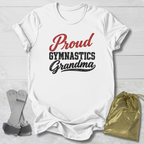 A white short-sleeve T-shirt laid flat on a light wood background, printed with the words 'Proud Gymnastics Grandma' in red and black lettering. A small gold drawstring pouch and a pair of gray straps are placed beside the shirt as styling props.