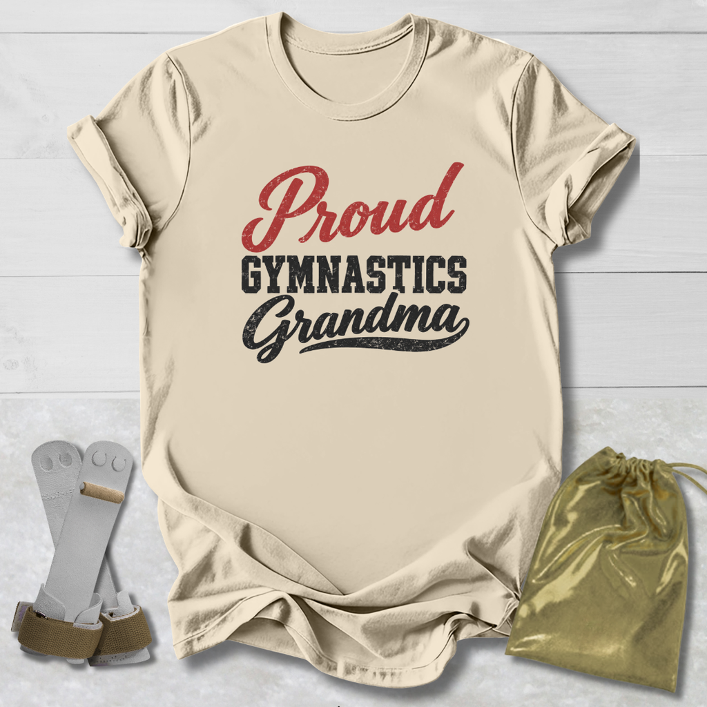 A beige short-sleeve T-shirt laid flat on a white wood background, printed with the words 'Proud Gymnastics Grandma' in red and black. A pair of gymnastics straps and a shiny gold drawstring pouch are placed beside the shirt as styling props.