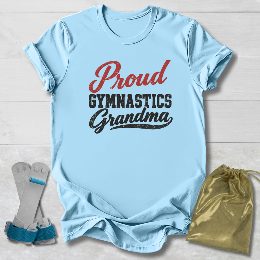 A light blue short-sleeve T-shirt laid flat on a white wood background, printed with the words 'Proud Gymnastics Grandma' in red and black. A pair of gymnastics straps and a shiny gold drawstring bag are placed beside the shirt as styling props.