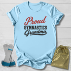 A light blue short-sleeve T-shirt laid flat on a white wood background, printed with the words 'Proud Gymnastics Grandma' in red and black. A pair of gymnastics straps and a shiny gold drawstring bag are placed beside the shirt as styling props.