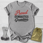 A heather gray short-sleeve T-shirt laid flat on a white wood background, printed with the words 'Proud Gymnastics Grandma' in red and black. A small gold drawstring pouch and a pair of gymnastics straps are placed beside the shirt as included accessories or styling props.
