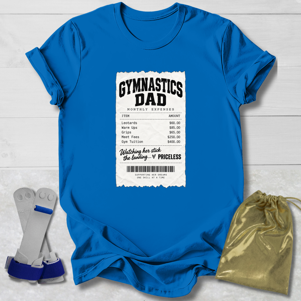 A bright blue short-sleeve t-shirt laid flat on a white wood background, featuring a white printed gymnasium ticket graphic on the front with the words 'GYMNASTICS DAD' and a list of expenses. A gold drawstring pouch and a pair of gray and blue gymnastics bars are placed beside the shirt.
