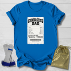 A bright blue short-sleeve t-shirt laid flat on a white wood background, featuring a white printed gymnasium ticket graphic on the front with the words 'GYMNASTICS DAD' and a list of expenses. A gold drawstring pouch and a pair of gray and blue gymnastics bars are placed beside the shirt.