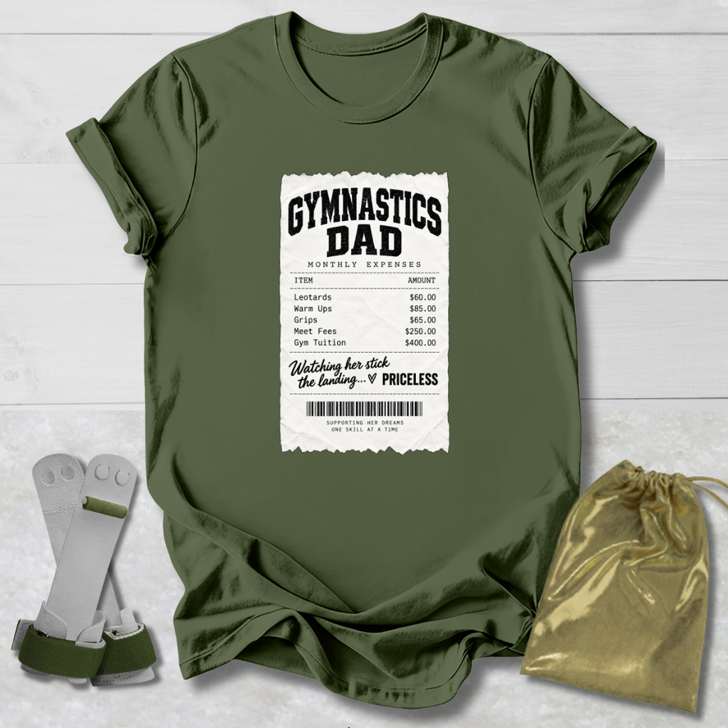An olive green short-sleeve t-shirt laid flat on a white wood background, featuring a large white graphic print that reads 'GYMNASTICS DAD' with a mock expense ticket design. A pair of gymnastics straps and a gold drawstring pouch are placed beside the shirt as styling props.