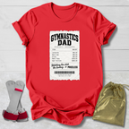 A red short-sleeve T-shirt laid flat on a light wood background, featuring a large white ticket-style graphic on the front with the text 'GYMNASTICS DAD' and a printed expense table. A pair of gymnastics trampoline bars and a gold drawstring pouch are placed beside the shirt as styling props.