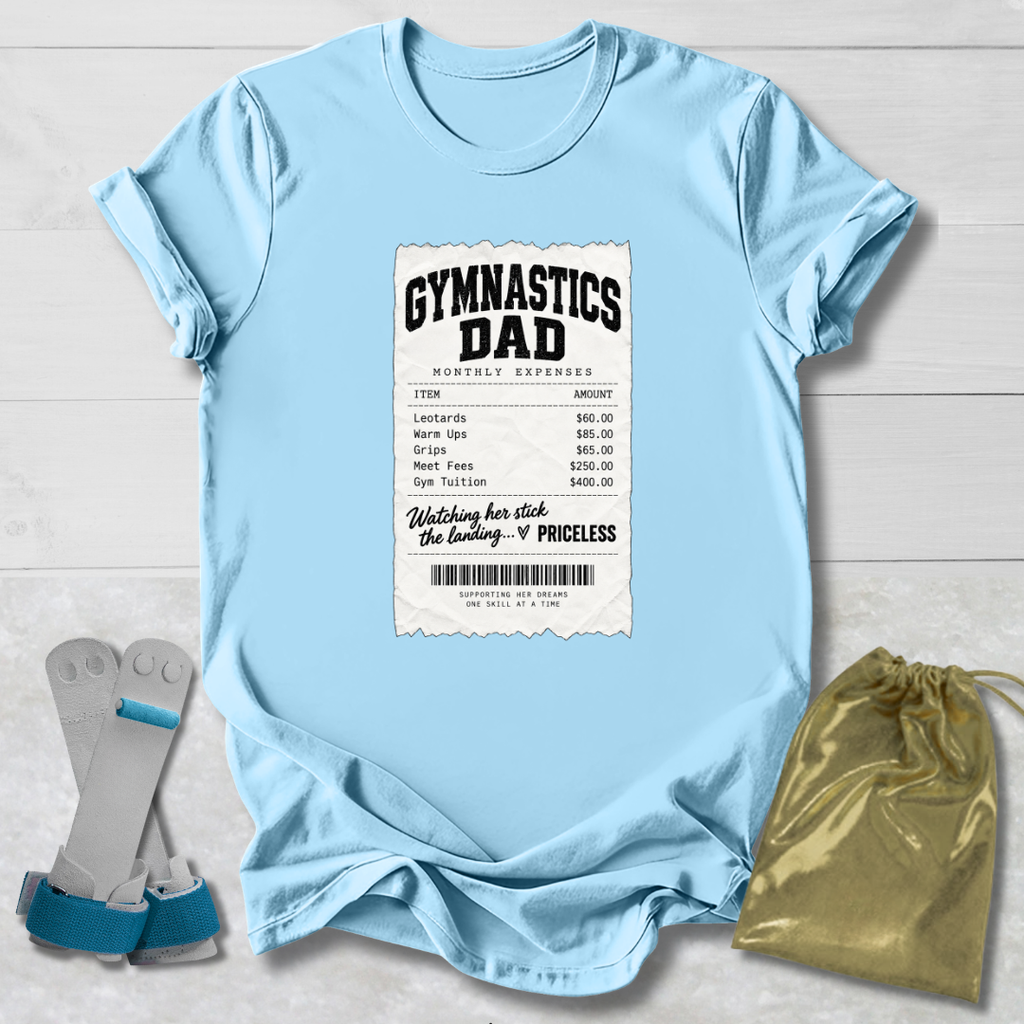 A light blue graphic t-shirt laid flat on a white wood background, featuring a printed gymnasium expense ticket design with the words 'GYMNASTICS DAD'. A pair of blue and gray gymnastics straps and a gold drawstring pouch are placed beside the shirt as styling props.