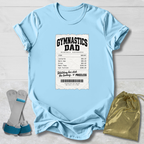 A light blue graphic t-shirt laid flat on a white wood background, featuring a printed gymnasium expense ticket design with the words 'GYMNASTICS DAD'. A pair of blue and gray gymnastics straps and a gold drawstring pouch are placed beside the shirt as styling props.
