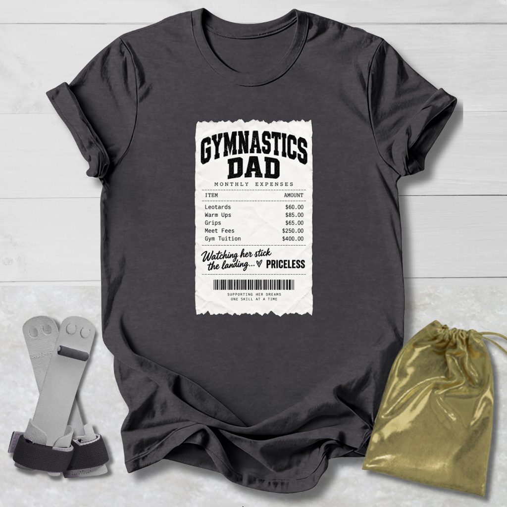 A dark charcoal gray graphic t-shirt laid flat on a light gray wood background, featuring a white and black 'Gymnastics Dad' receipt-style print on the front. A gold drawstring pouch and a pair of gray resistance bands are placed beside the shirt as styling props.