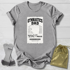 A heather gray short-sleeve t-shirt laid flat on a light wood background, featuring a white printed ticket graphic that reads 'GYMNASTICS DAD' with a billing table and barcode. A gray adjustable strap accessory, a gold drawstring pouch, and a small gray item are placed around the shirt as styling props.