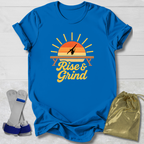 A bright blue graphic T-shirt laid flat on a white wood background, featuring a retro sunset design with a gymnast silhouette and the words 'Rise & Grind.' Two gray-and-blue wrist wraps and a metallic gold drawstring pouch are placed beside the shirt as included accessories.