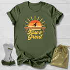 An olive green graphic t-shirt laid flat on a white wood background, featuring a retro sunset design with a gymnast silhouette and the words 'Rise & Grind.' Two small accessories are placed beside it: a pair of white and green straps on the left and a shiny gold drawstring pouch on the right.