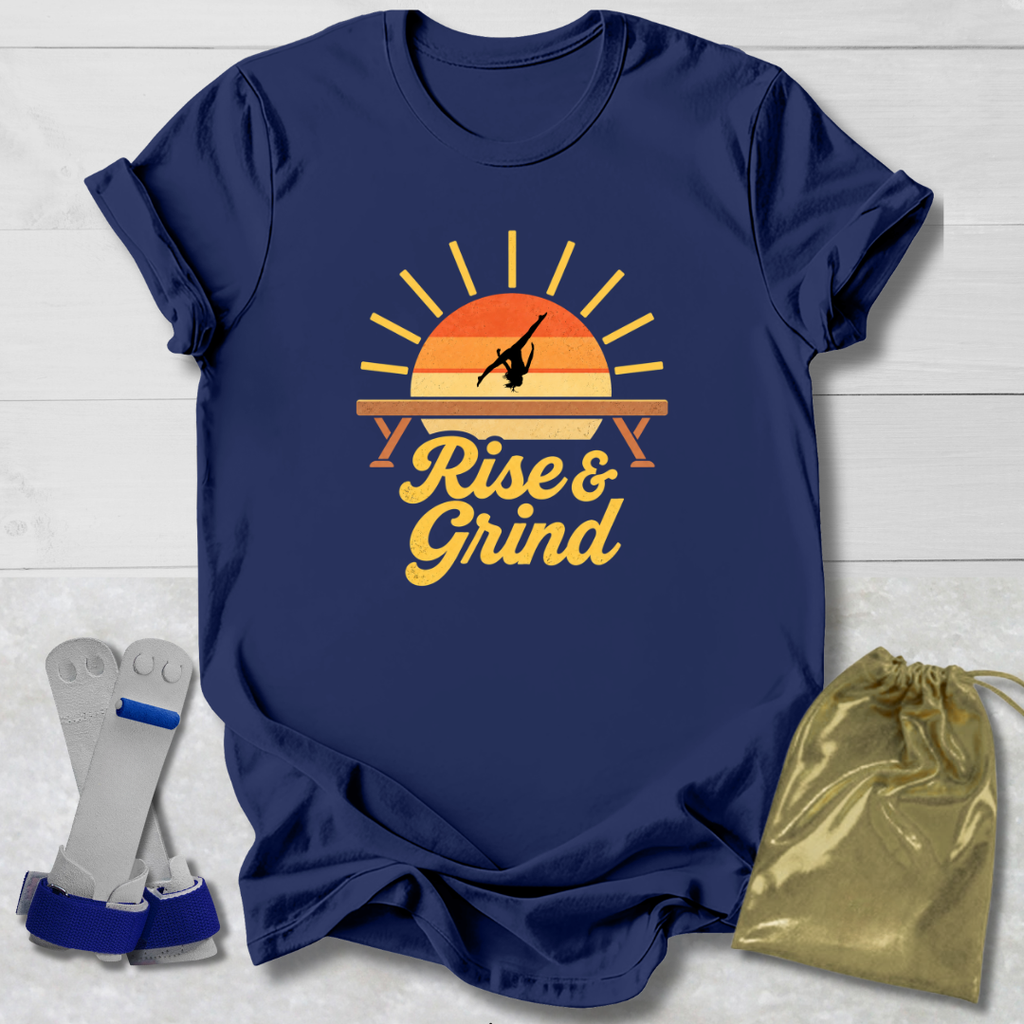 A navy blue graphic t-shirt laid flat on a white wood background, featuring a retro sunset design with the words 'Rise & Grind.' Two gray and blue weight plates are placed to the left, and a shiny gold drawstring bag is to the right, suggesting a workout bundle.