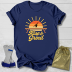 A navy blue graphic t-shirt laid flat on a white wood background, featuring a retro sunset design with the words 'Rise & Grind.' Two gray and blue weight plates are placed to the left, and a shiny gold drawstring bag is to the right, suggesting a workout bundle.