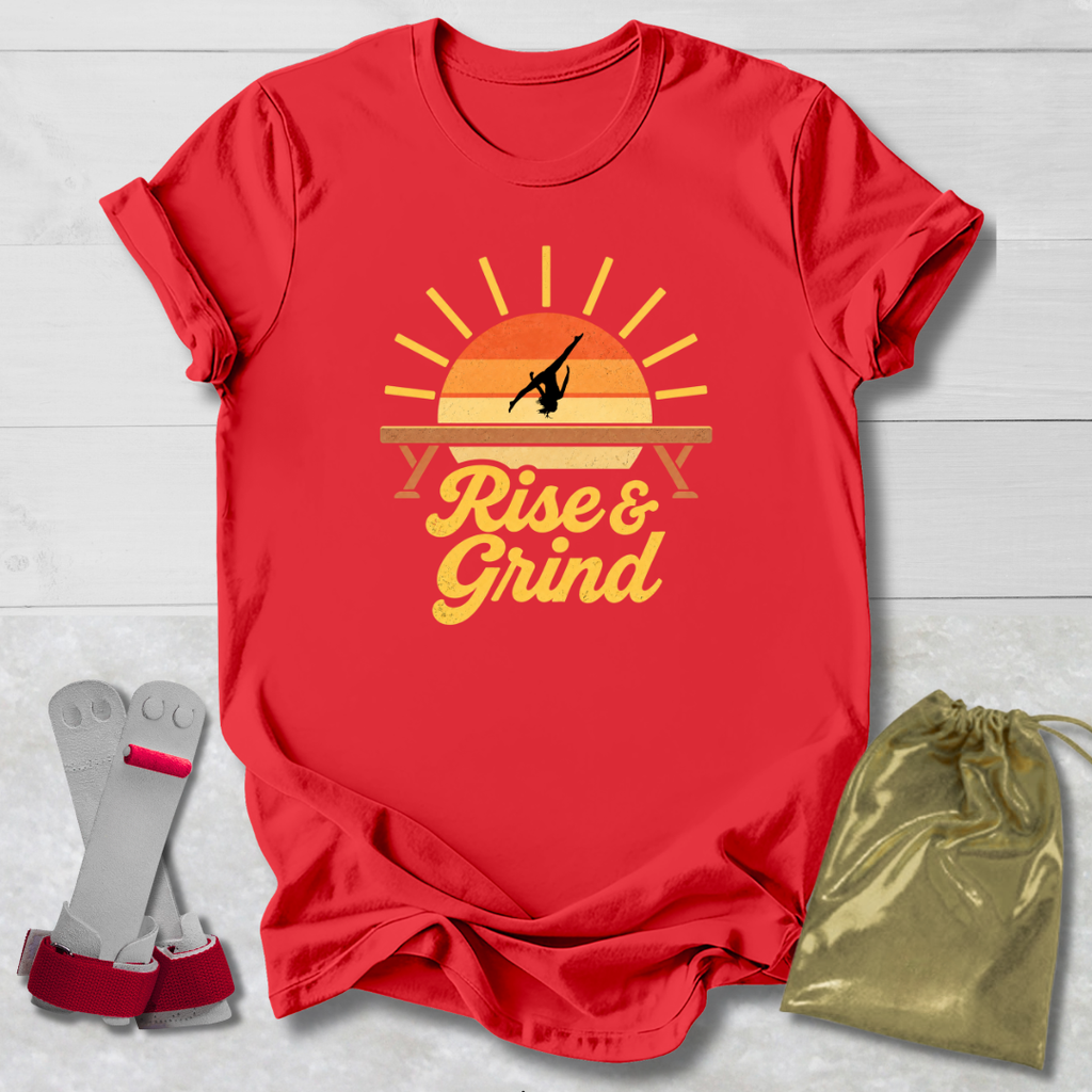 A red short-sleeve graphic T-shirt laid flat on a light wood background, featuring a retro sunset design with a gymnast silhouette and the words 'Rise & Grind.' Two gray resistance bands with pink bands are placed to the left, and a shiny gold drawstring pouch is placed to the right.