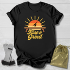 A black short-sleeve graphic T-shirt laid flat on a light wood background, featuring a retro sunset-style print with the words 'Rise & Grind' in yellow. A gold drawstring pouch and a pair of gray wrist wraps are placed beside the shirt as styling props.