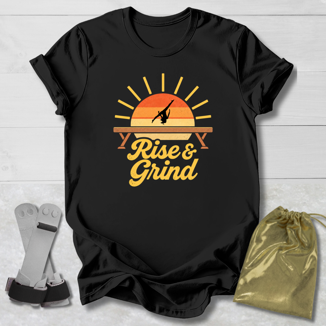 A black short-sleeve graphic T-shirt laid flat on a light wood background, featuring a retro sunset-style print with the words 'Rise & Grind' in yellow. A gold drawstring pouch and a pair of gray wrist wraps are placed beside the shirt as styling props.