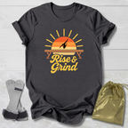 A dark charcoal gray t-shirt with a retro sunset graphic and the words 'Rise & Grind' in yellow, styled in a flat-lay with a gold drawstring bag and gray wrist wraps on a white wood background.