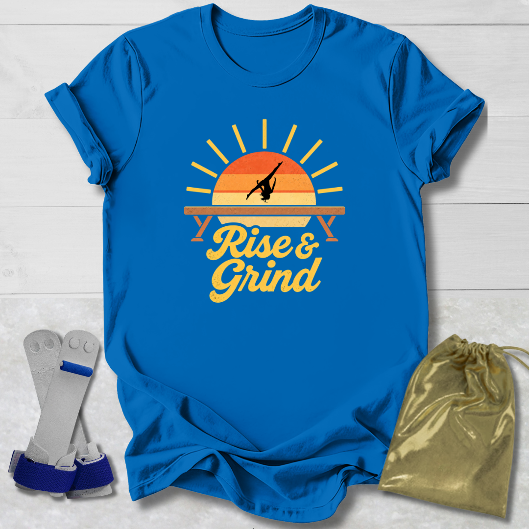 A bright blue graphic T-shirt laid flat on a white wood background, featuring a retro sunset design with a gymnast silhouette and the words 'Rise & Grind.' Two gray-and-blue wrist wraps and a metallic gold drawstring pouch are placed beside the shirt as included accessories.