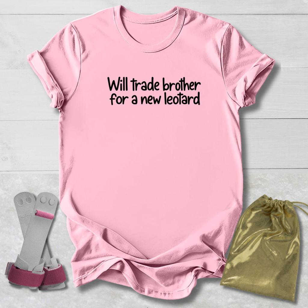 A light pink short-sleeve t-shirt with the black text 'Will trade brother for a new leopard' printed on the chest, displayed on a light gray wood background alongside a pair of gray and pink boxing gloves and a gold drawstring bag.