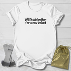 A white short-sleeve T-shirt laid flat on a light wood background, printed with the black text 'Will trade brother for a new leopard' across the chest. Two small accessories are placed beside it: a pair of gray straps on the left and a shiny gold drawstring pouch on the right.